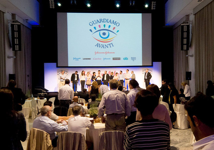 Convention guardiamo avanti, il pubblico di spalle guarda i membri sul palco sotto il logo aziendale proiettato sullo schermo