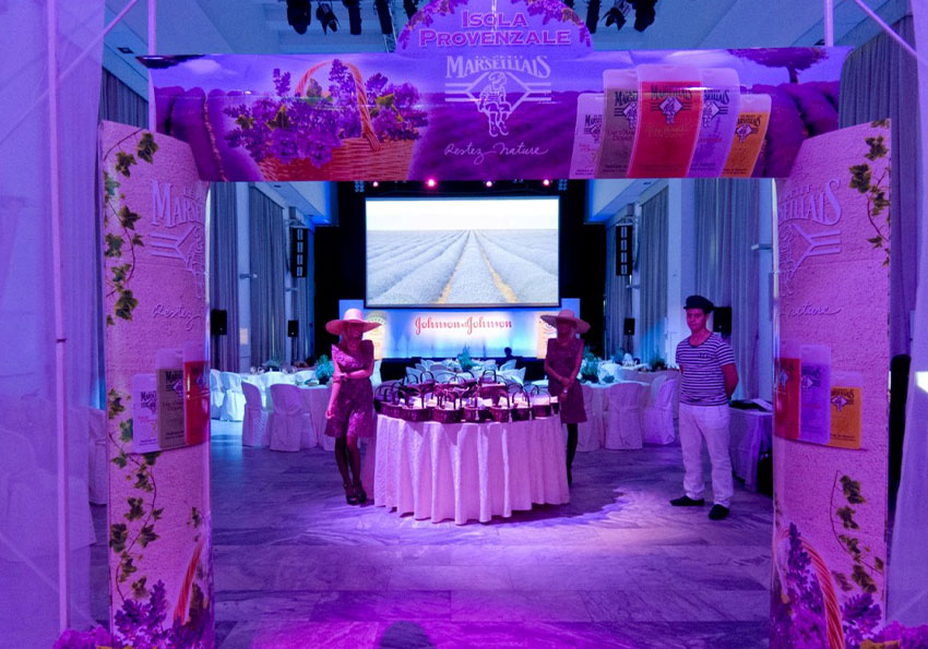 Lancio prodotto isola provenzale con arco d'ingresso alla sala personalizzato, un tavolo circolare allestito, due hostess e uno stuard vestiti in stile provenzale